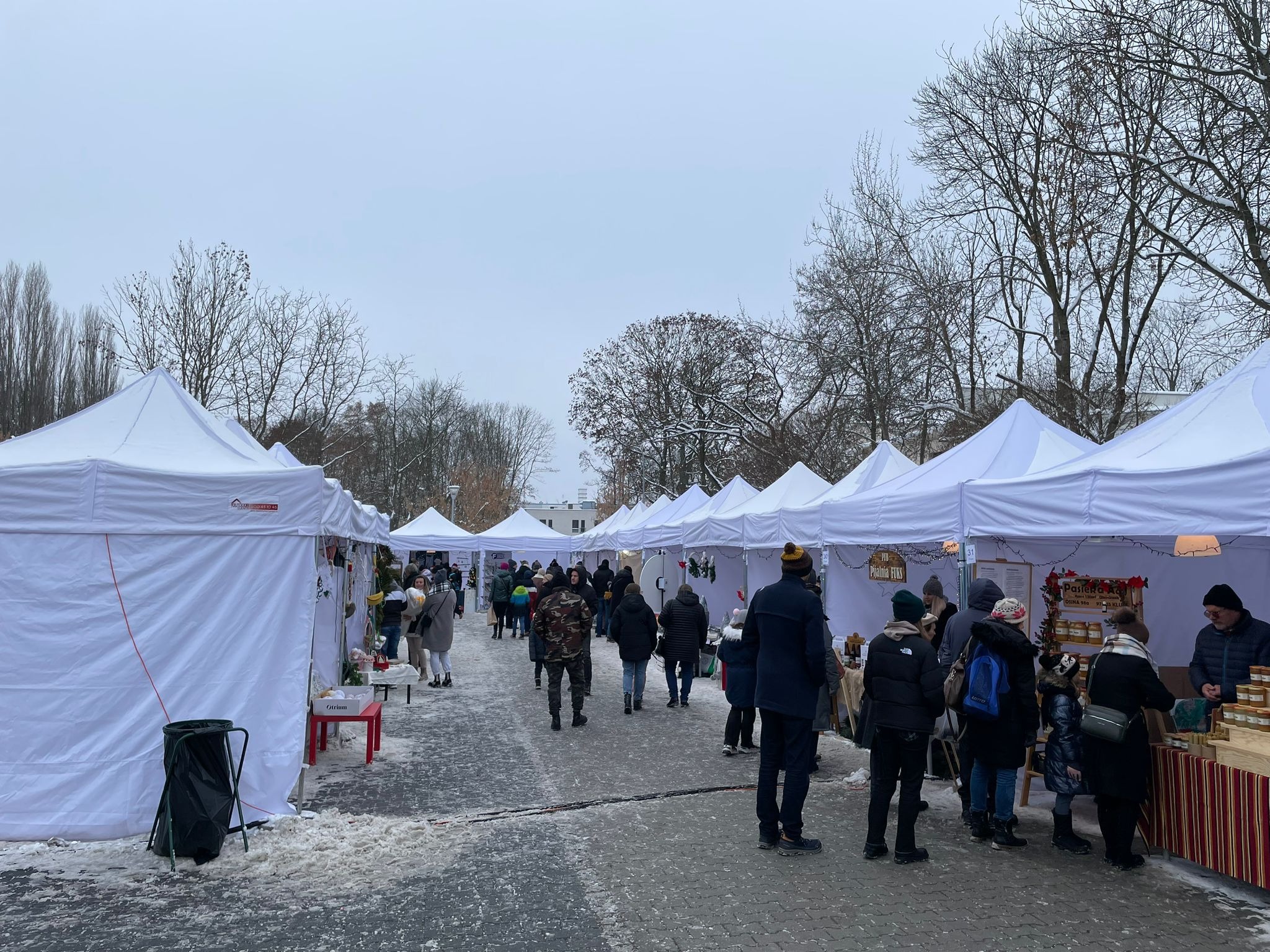 Zimowy jarmark na zewnątrz z rzędem białych namiotów, straganów i spacerujących ludzi w zimowych ubraniach, śnieg na ziemi.