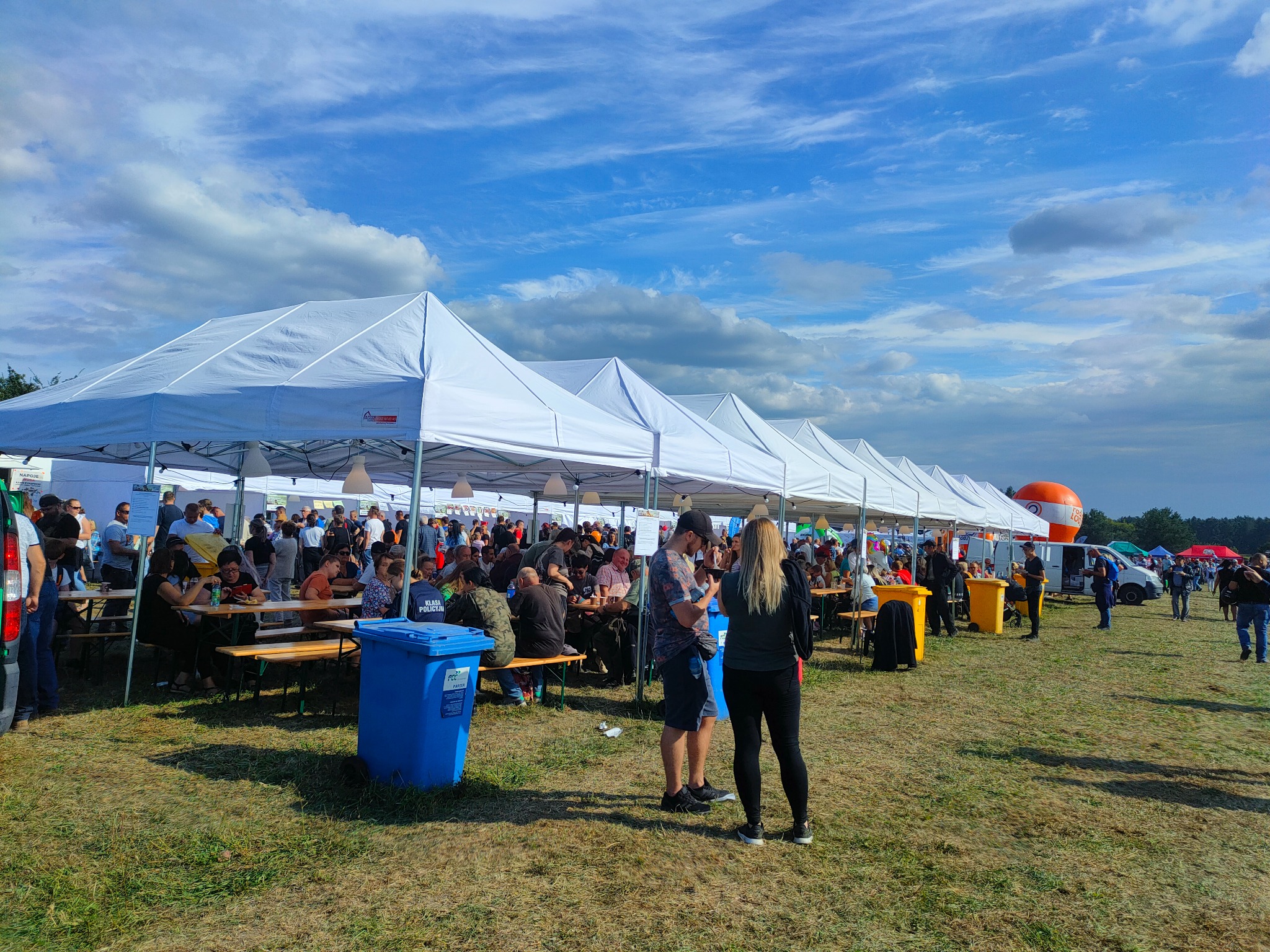 Długi rząd białych namiotów cateringowych pod błękitnym niebem z chmurami, ustawionych na trawiastym terenie, z ludźmi siedzącymi przy stołach i koszami na śmieci w kolorze niebieskim i żółtym.