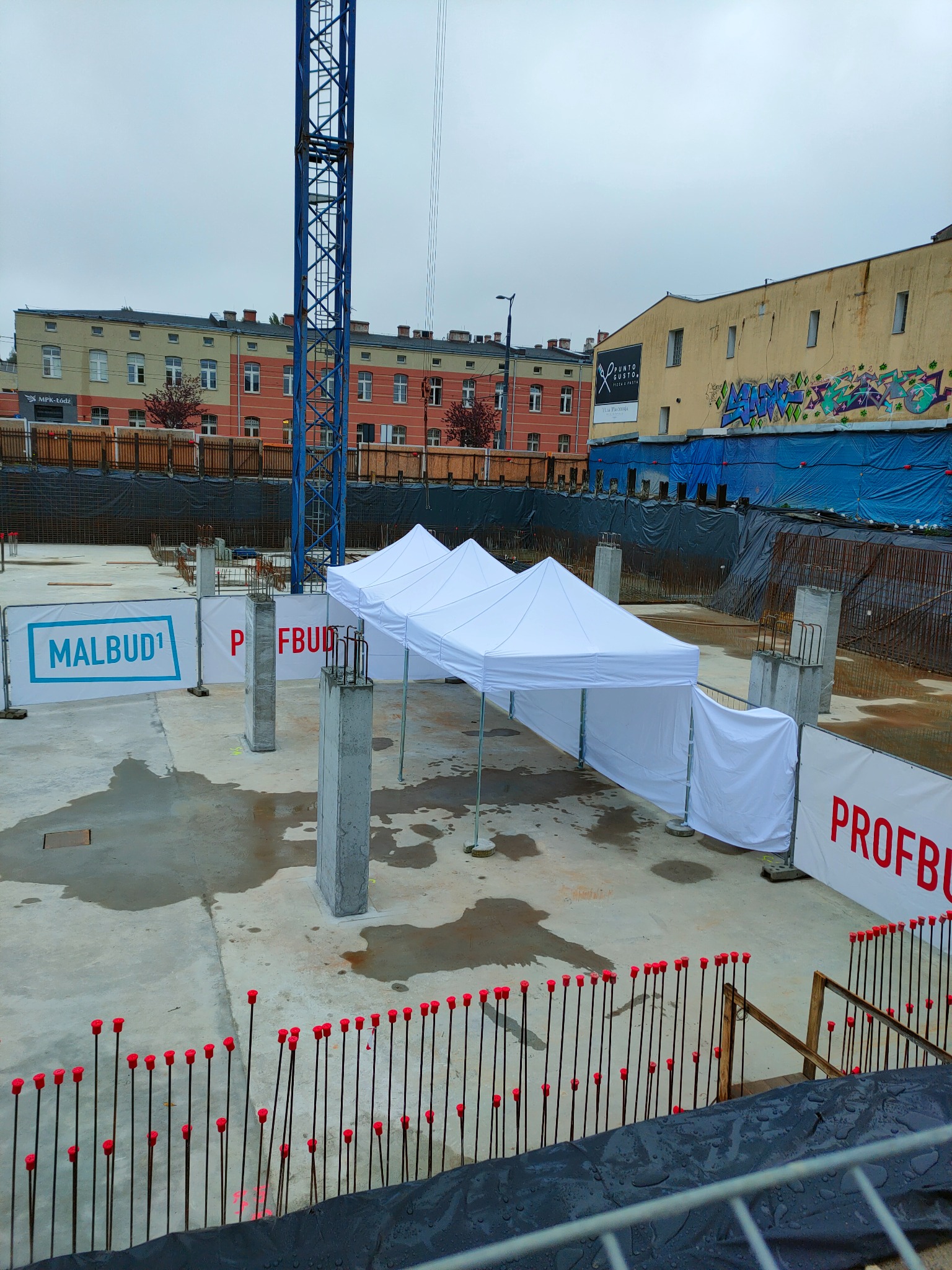 Biały namiot rozstawiony na placu budowy z widocznymi elementami konstrukcyjnymi i tablicami informacyjnymi firm budowlanych w tle.