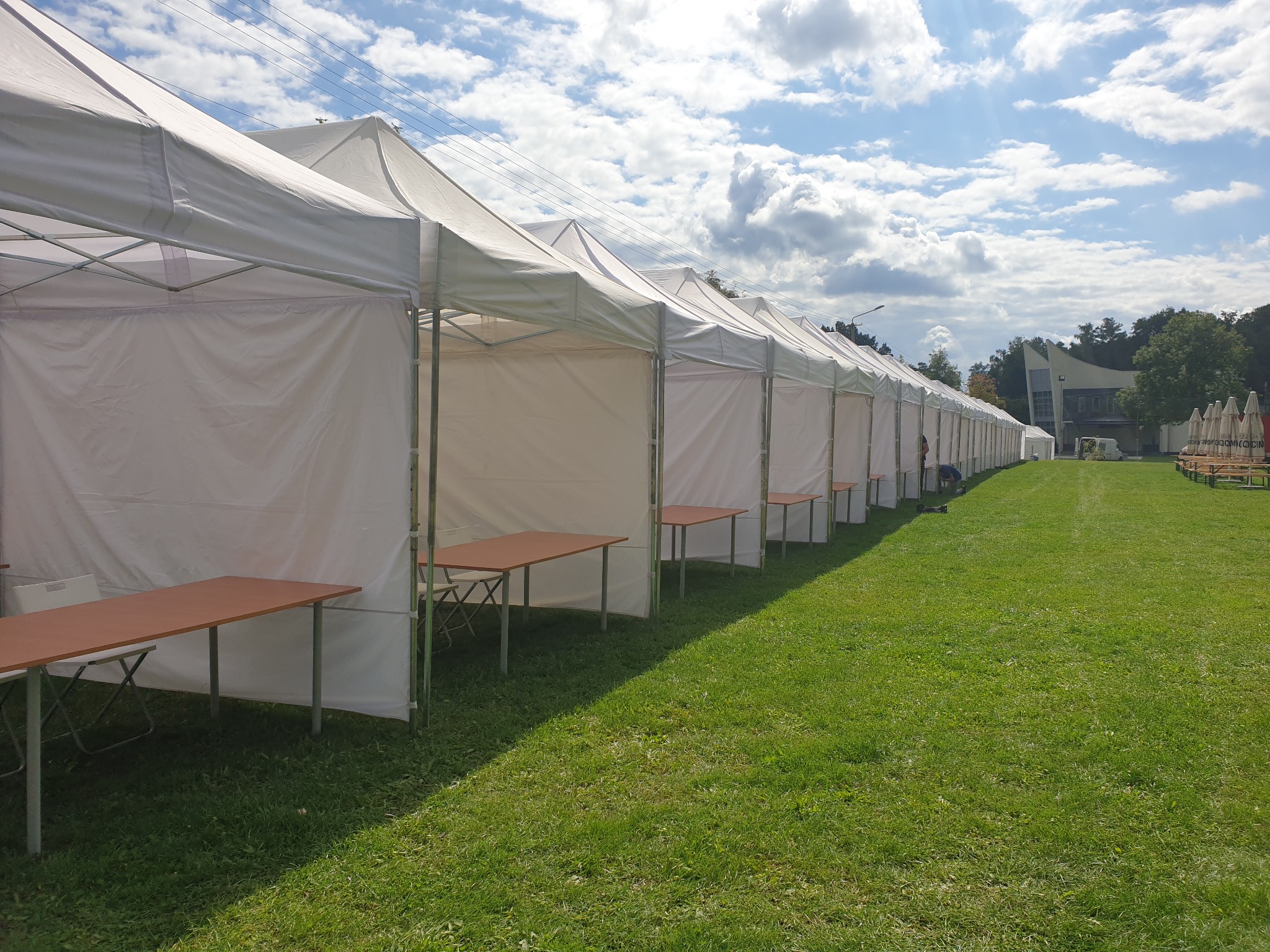 Rząd białych namiotów eventowych z brązowymi stołami na zielonej trawie, błękitne niebo z chmurami w tle.