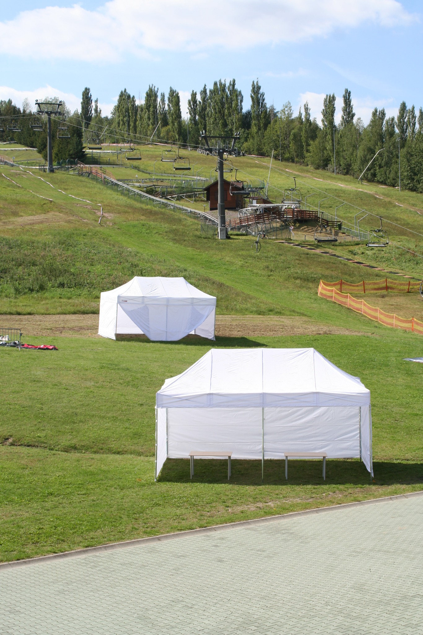 Dwa białe namioty ustawione na zielonej trawie, w tle wyciąg narciarski i drzewa pod błękitnym niebem.
