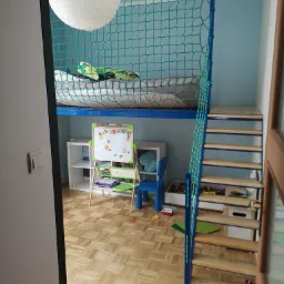 Pokój dziecięcy z łóżkiem na antresoli zabezpieczonym siatką, dostępnym po drewnianych schodach z niebieskimi elementami, pod łóżkiem biurko i regał na zabawki.