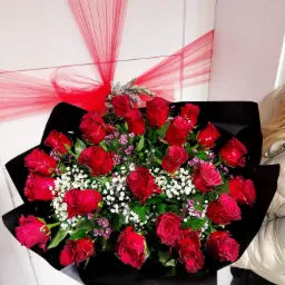 Duży bukiet czerwonych róż z białymi i fioletowymi drobnymi kwiatami, owinięty w czarny papier, z dekoracją z czerwonej tiulowej wstążki, na tle białych drzwi z napisem 'Pomieszczenie Socjalne'.