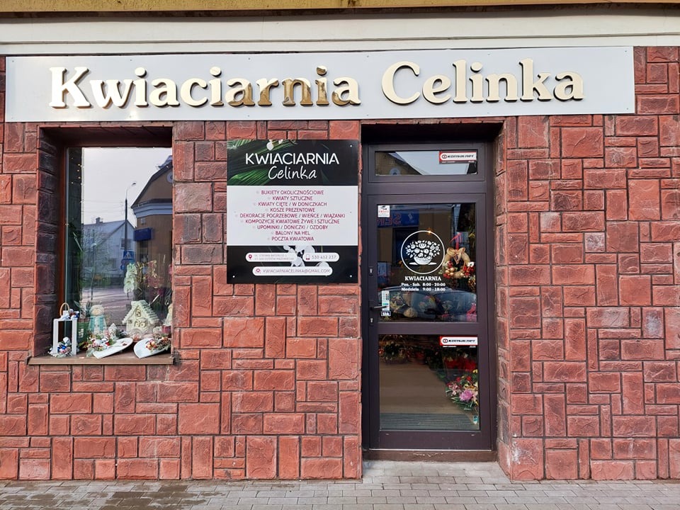 Fasada kwiaciarni Celinka z cegły z witryną okienną prezentującą dekoracje i szyldem z nazwą. Na ścianie tablica informacyjna z ofertą i danymi kontaktowymi.