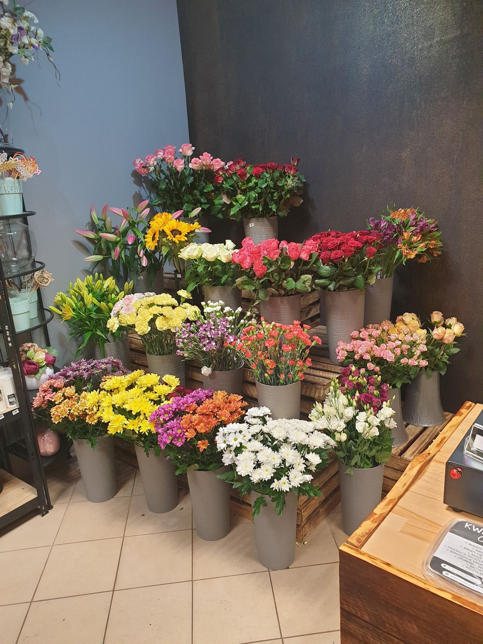 Ekspozycja różnorodnych kwiatów w szarych, cylindrycznych wazonach, ustawionych na drewnianych skrzynkach, tworzących schodkową kompozycję na tle ciemnej ściany w kwiaciarni.