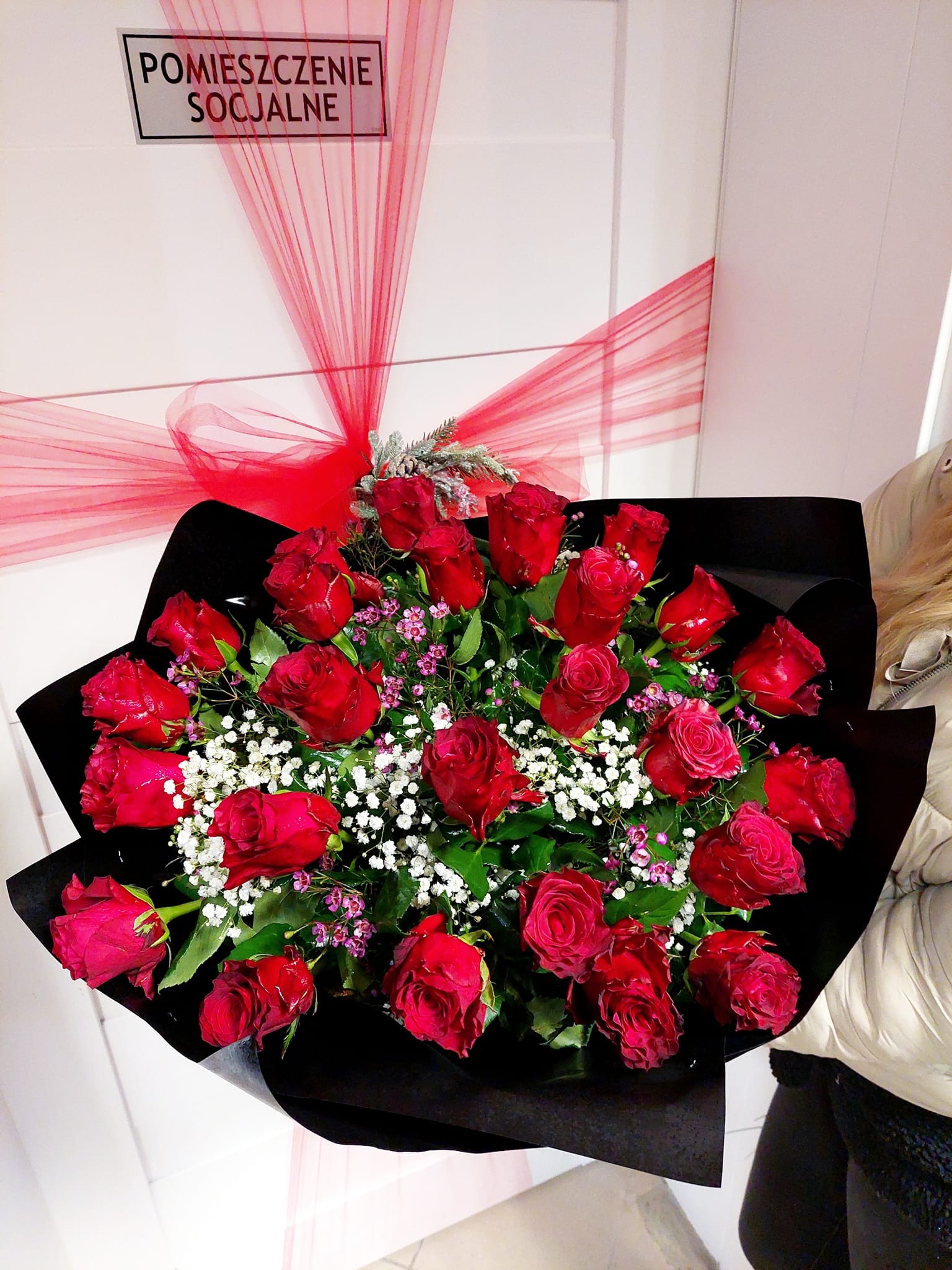 Duży bukiet czerwonych róż z białymi i fioletowymi drobnymi kwiatami, owinięty w czarny papier, z dekoracją z czerwonej tiulowej wstążki, na tle białych drzwi z napisem 'Pomieszczenie Socjalne'.