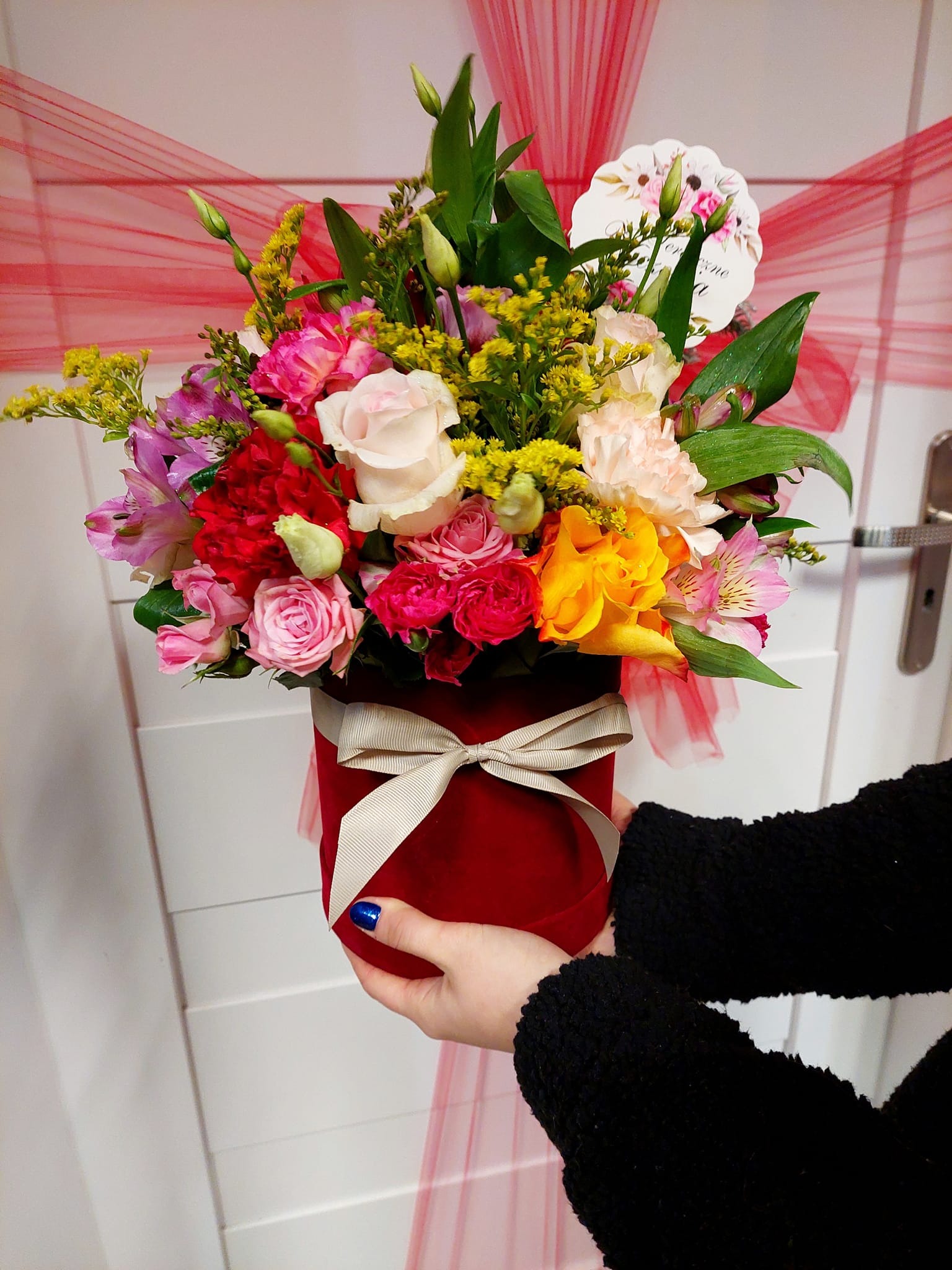 Ręce trzymają czerwony, okrągły pudełko z mieszanym bukietem róż, alstromerii i żółtych kwiatów, przewiązane kremową wstążką, na tle białych drzwi z czerwoną, prześwitującą tkaniną.