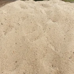 Duża pryzma przesiewanego piasku budowlanego o drobnej granulacji, z widocznymi drobnymi ciemnymi kamyczkami i źdźbłami trawy, oświetlona naturalnym światłem słonecznym.