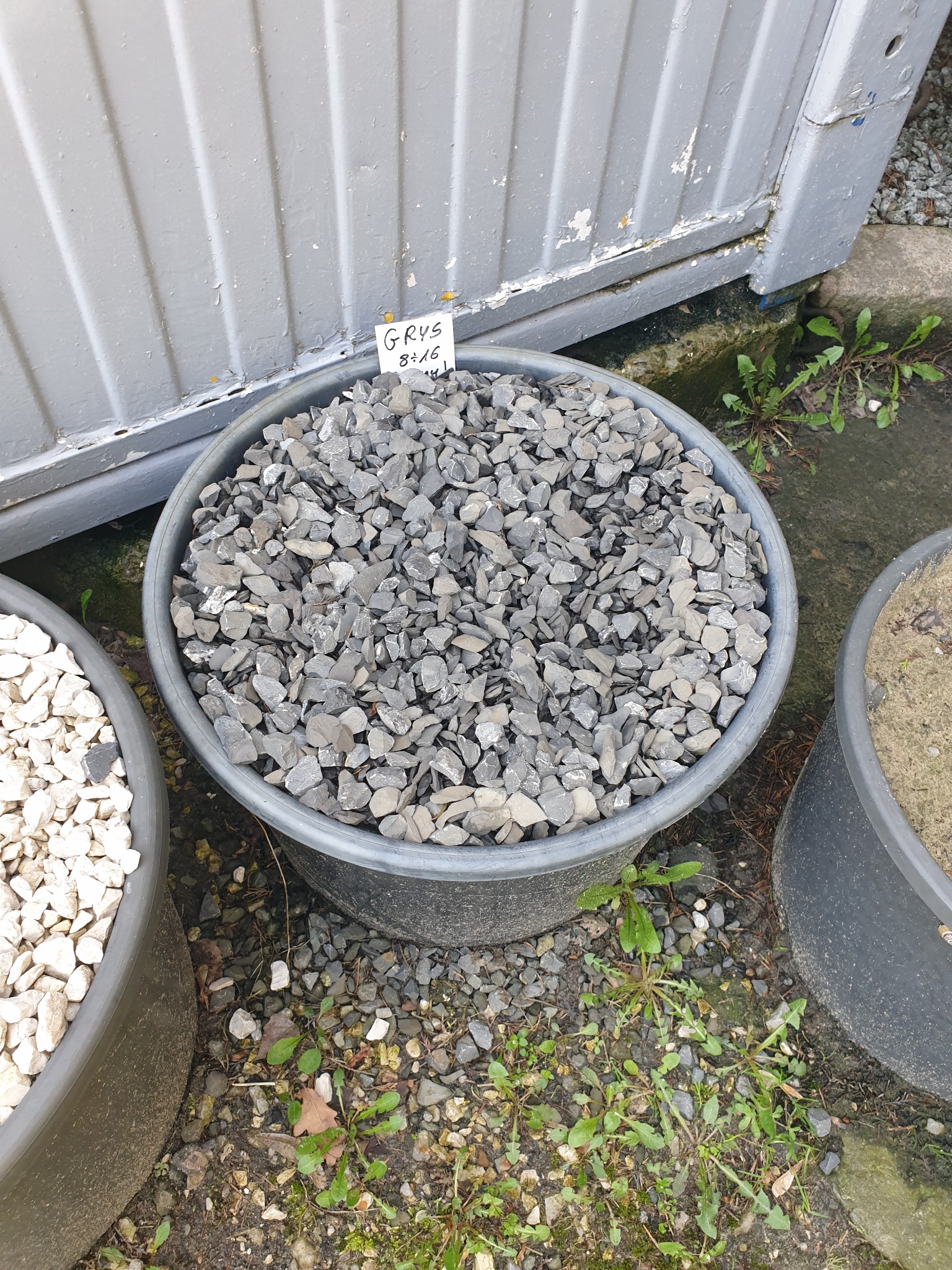 Grys 8-16mm w czarnym plastikowym pojemniku, obok inne pojemniki z białym kamieniem i piaskiem, na tle szarej, łuszczącej się ściany.