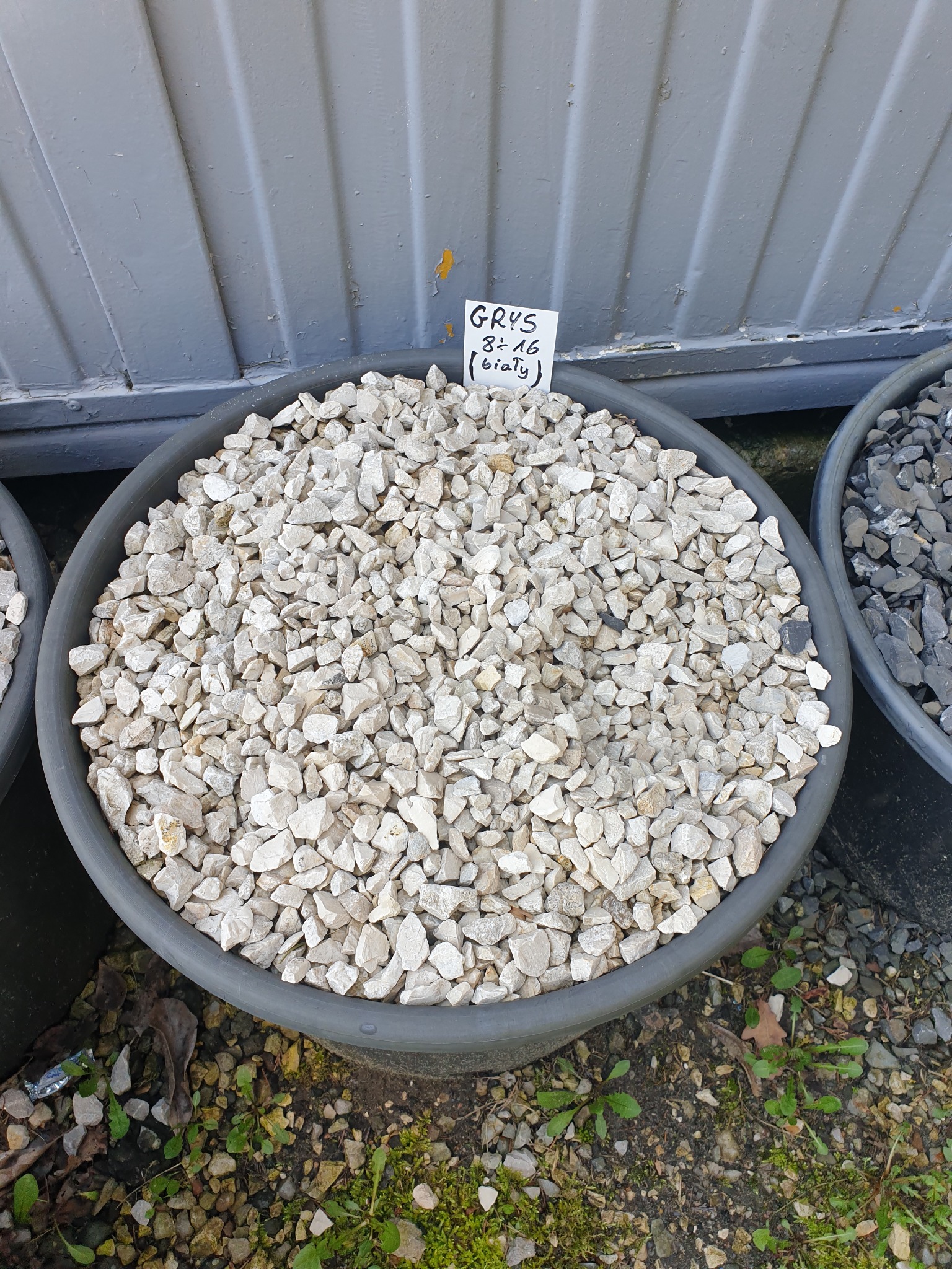 Biały grys w szarej donicy z kartką informacyjną, widoczne inne donice i podłoże z kamykami i roślinami.