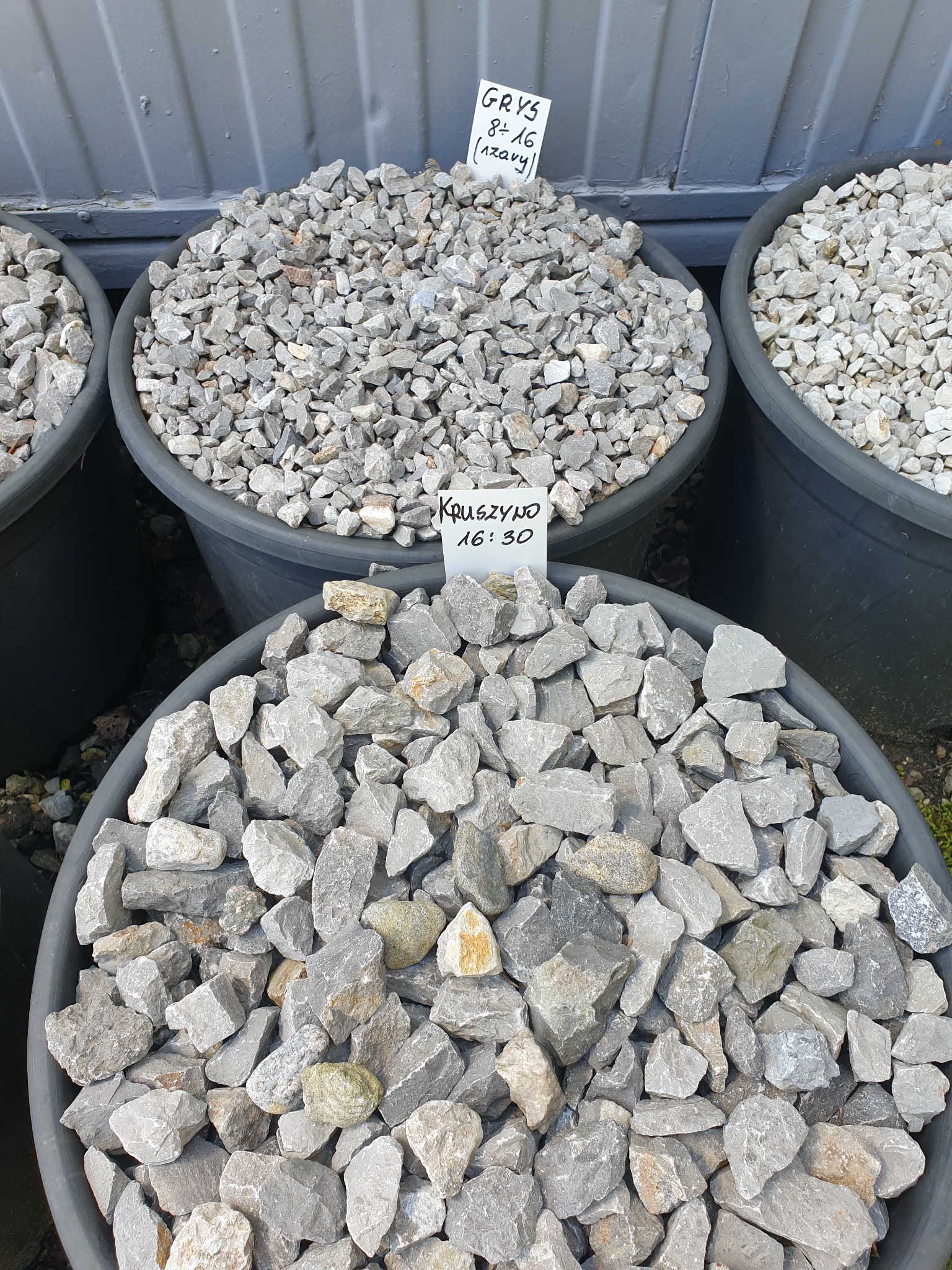 Grys i kruszywo w czarnych donicach, z widocznymi kartkami z cenami i informacjami o produkcie, ustawione na zewnątrz.