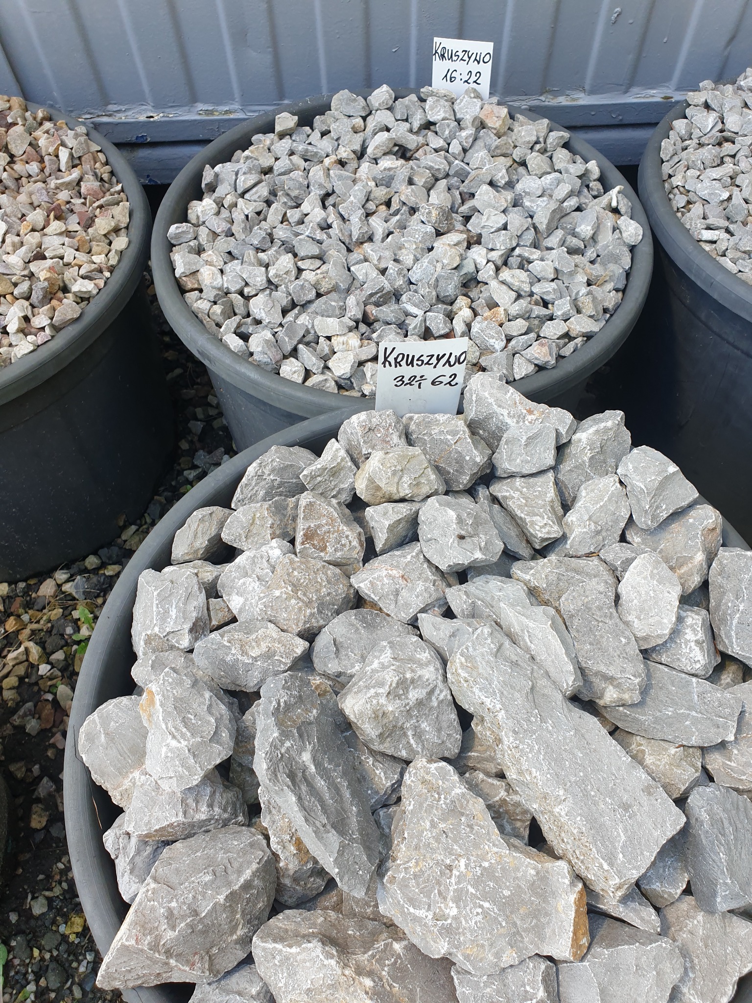 Kruszywo w trzech czarnych pojemnikach, z tabliczkami informacyjnymi z cenami. Większe, szare kamienie w pierwszym planie.