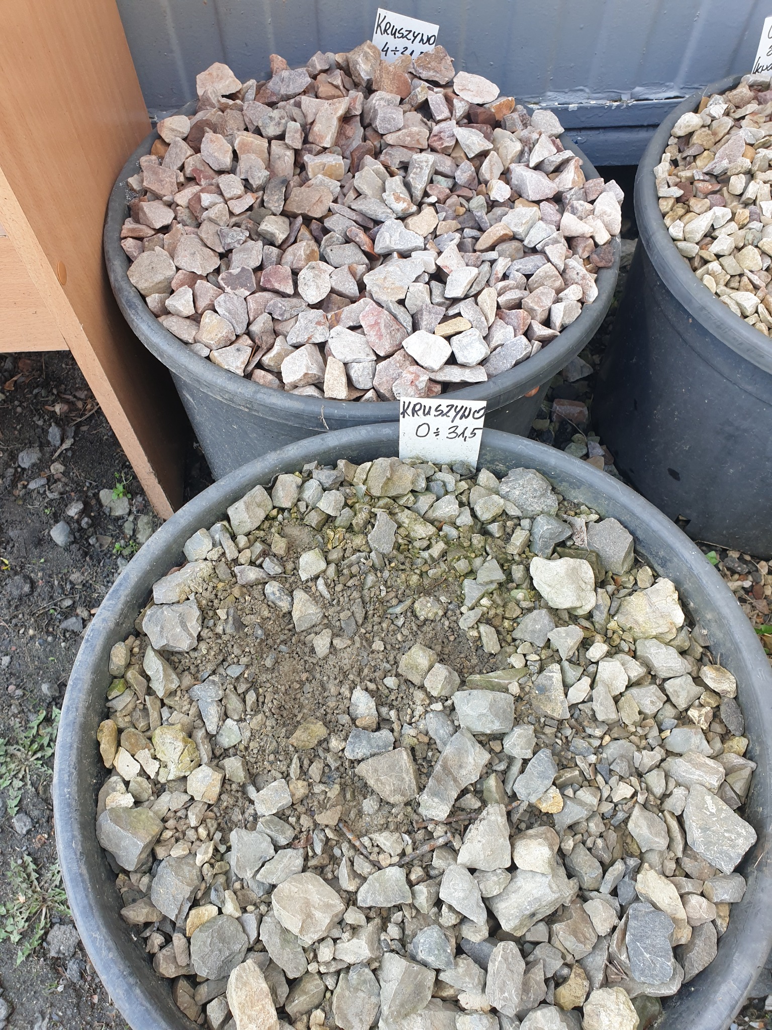 Kruszywo w plastikowych pojemnikach z etykietami informacyjnymi, prezentowane na zewnątrz, widoczne różne frakcje i kolory.