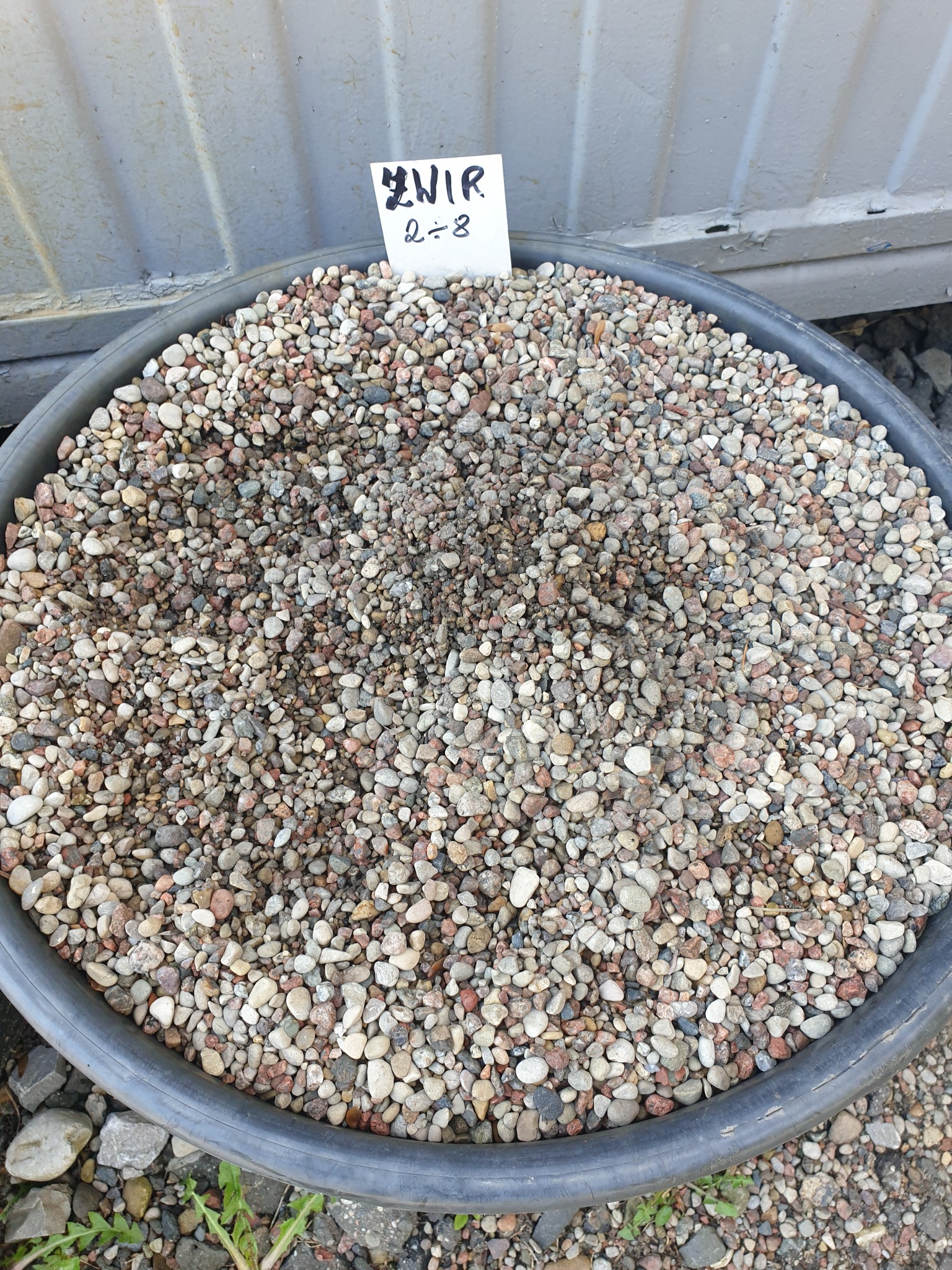 Okrągły, czarny pojemnik wypełniony drobnymi kamykami o zróżnicowanej kolorystyce, z widoczną kartką z napisem 'Żwir 2÷8' umieszczoną na wierzchu, na tle szarej, metalowej powierzchni.