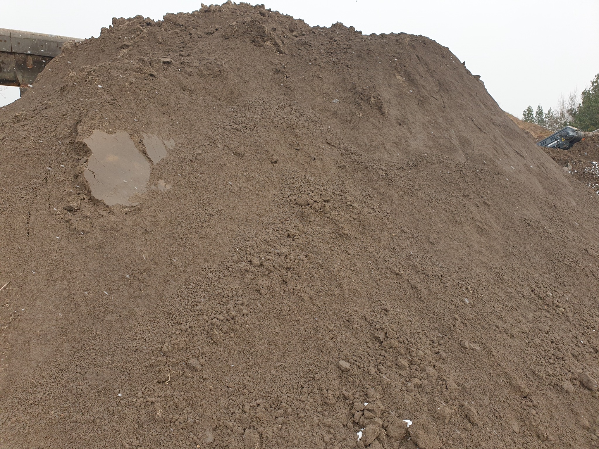 Duża pryzma brązowej ziemi ogrodowej z widocznym mokrym fragmentem, w tle fragment konstrukcji i drzewa.