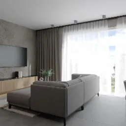 Nowoczesny salon z szarą sofą modułową, betonową ścianą z telewizorem i drewnianą szafką RTV, oświetlony światłem dziennym wpadającym przez okno z przezroczystymi zasłonami.