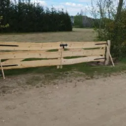 Surowa, drewniana brama wjazdowa z czarnymi zawiasami i prostym zamkiem, widoczna na tle zieleni i błękitnego nieba.