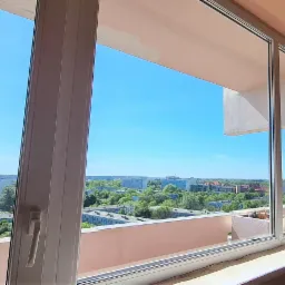 Widok z okna na miasto w słoneczny dzień, duże okno z białymi ramami, widoczny fragment balkonu i budynki w oddali.