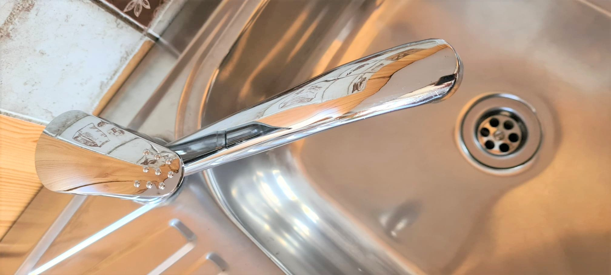 Błyszcząca, chromowana bateria umywalkowa z drobnymi otworami na perlator, ustawiona nad czystym, metalowym zlewem z widocznym odpływem.