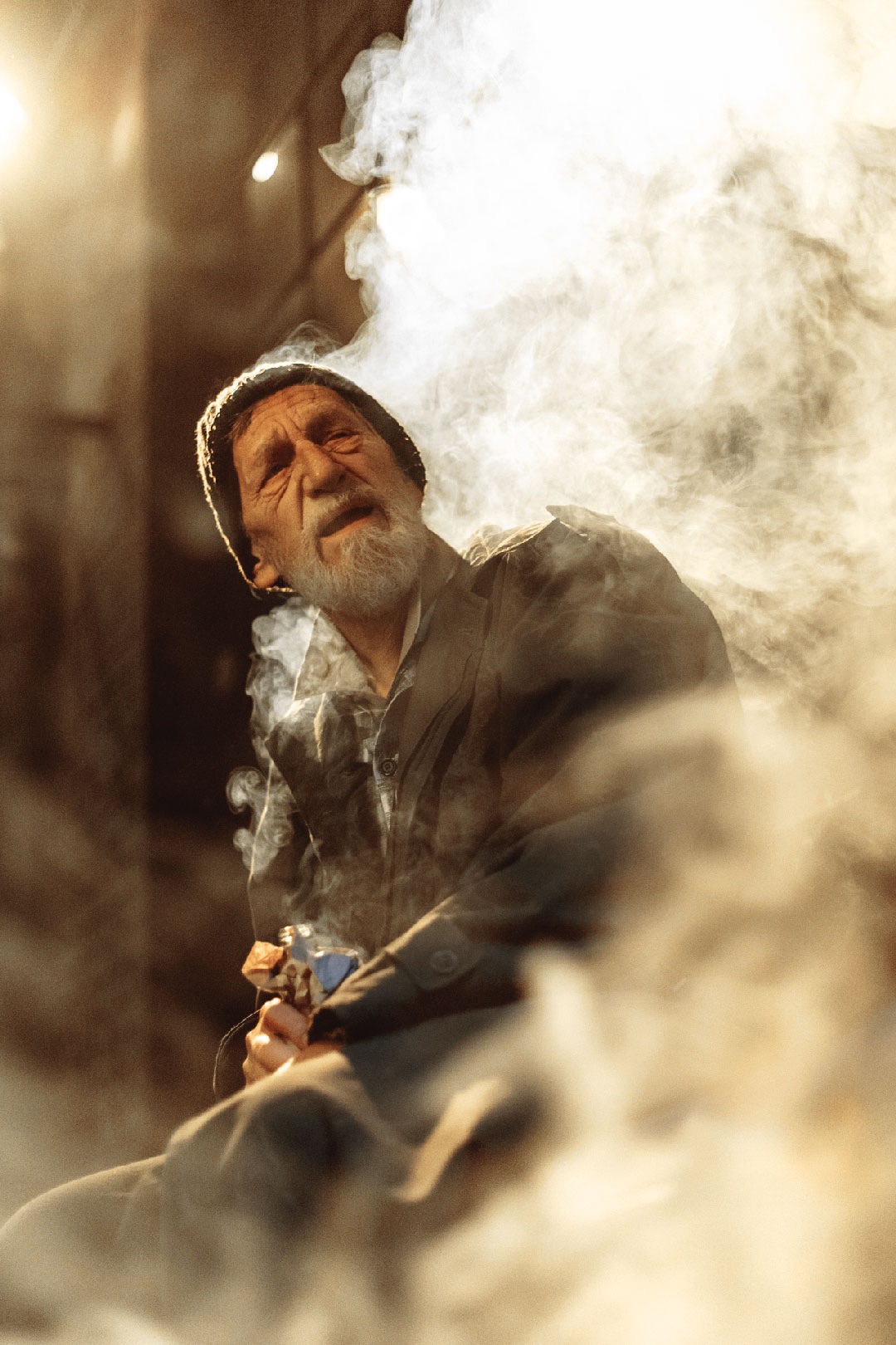Mężczyzna z brodą i czapce, oświetlony ciepłym światłem, w otoczeniu dymu, prawdopodobnie na planie filmowym.