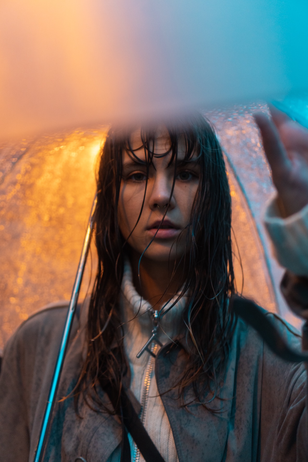 Portret kobiety pod przezroczystym parasolem w deszczu, oświetlonej pomarańczowym i niebieskim światłem, z mokrymi włosami opadającymi na twarz.