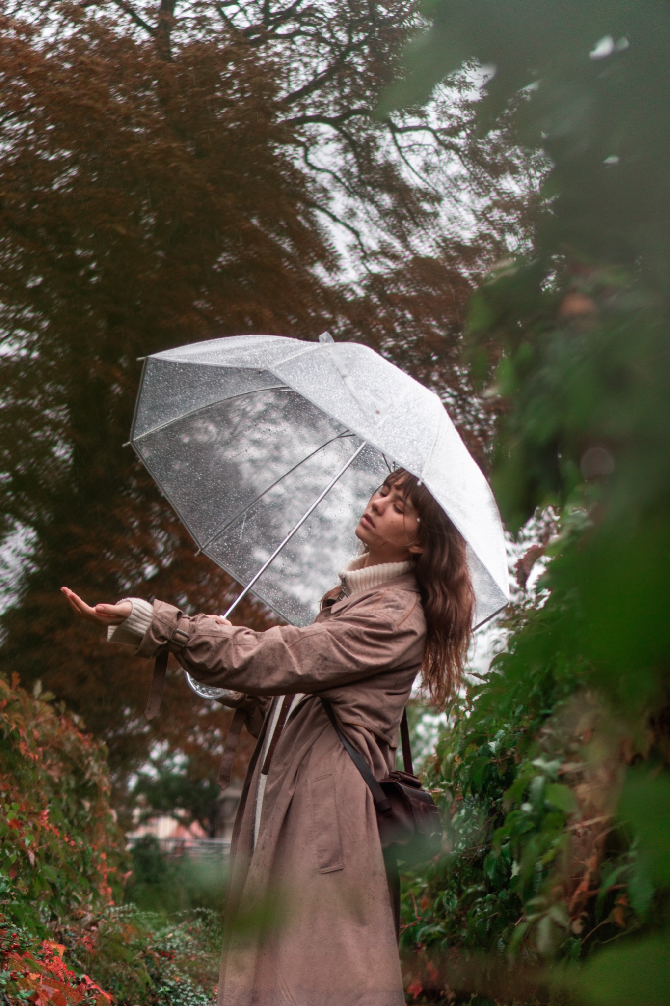Kobieta w płaszczu z przezroczystym parasolem, wyciągniętą dłonią czuje deszcz, jesienna sceneria z drzewami w tle.