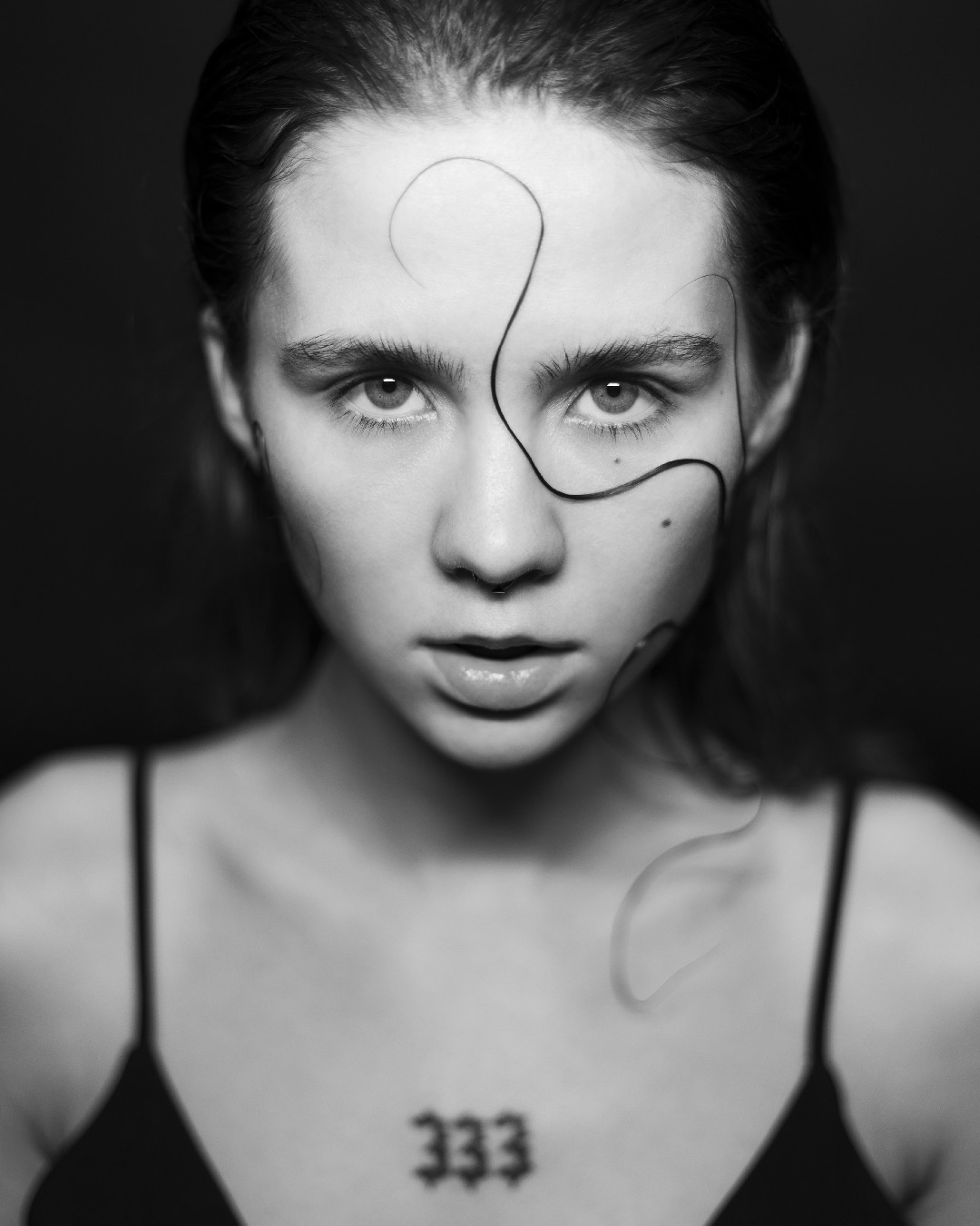Studyjny portret w czerni i bieli młodej kobiety z mokrymi włosami ułożonymi w abstrakcyjne wzory na twarzy, z tatuażem '333' na dekolcie, oświetlony z przodu.