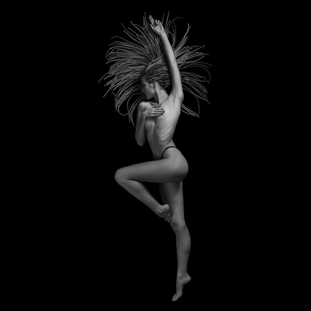 Czarno-białe zdjęcie studyjne: kobieta w dynamicznej pozie, z uniesioną ręką i długimi warkoczykami, na czarnym tle.