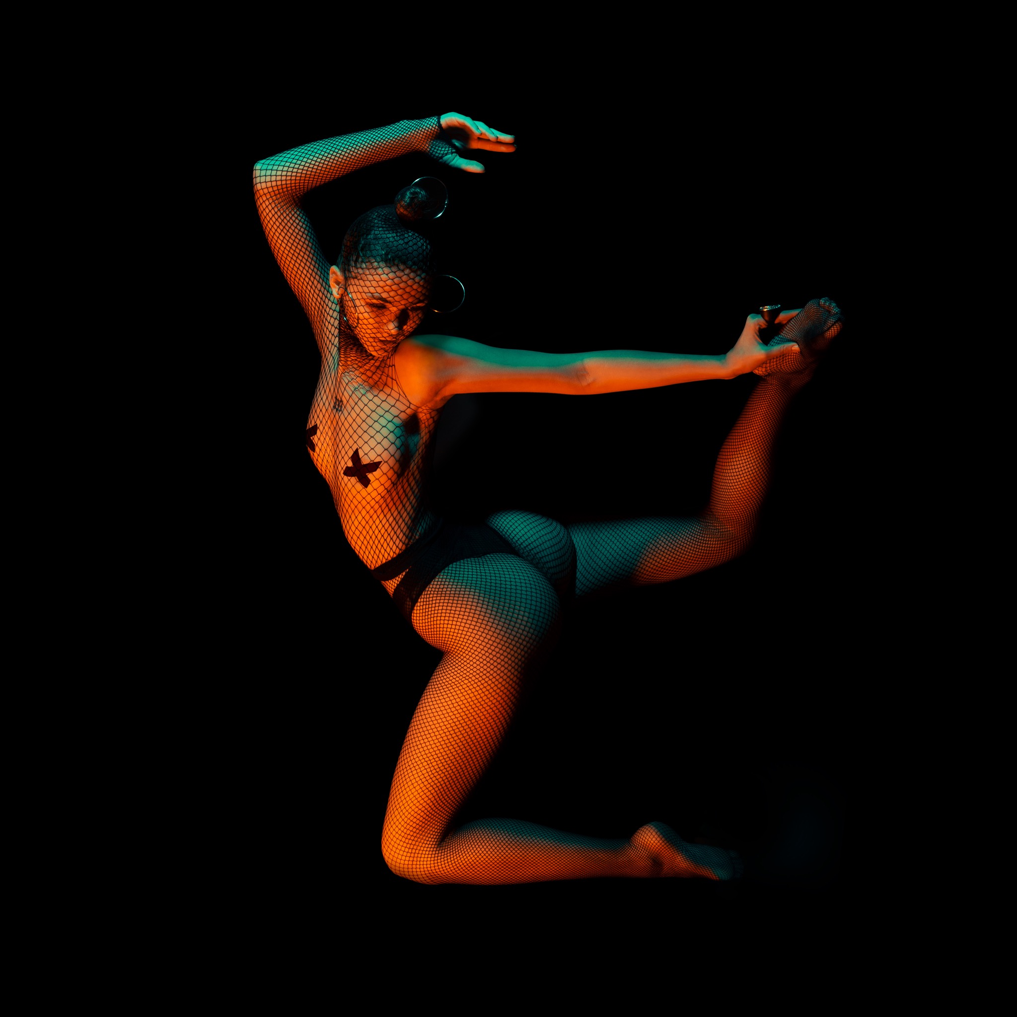 Studyjna sesja indywidualna: Kobieta w czarnej bieliźnie i kabaretkach, oświetlona pomarańczowym i turkusowym światłem, wykonuje taneczną pozę na czarnym tle.