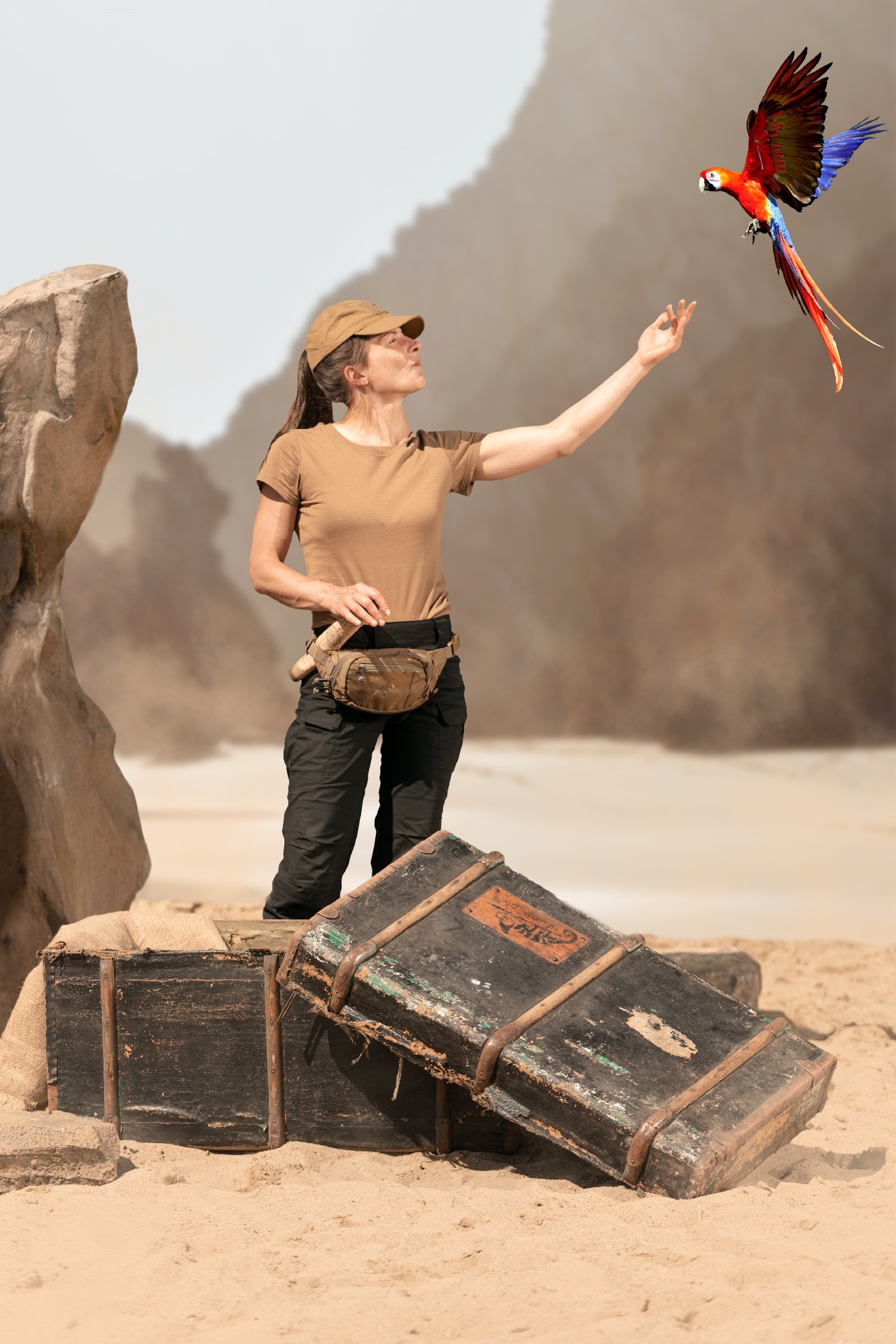 Kobieta w plenerze wyciąga rękę do lecącej papugi ara, obok leżą stare, drewniane skrzynie.