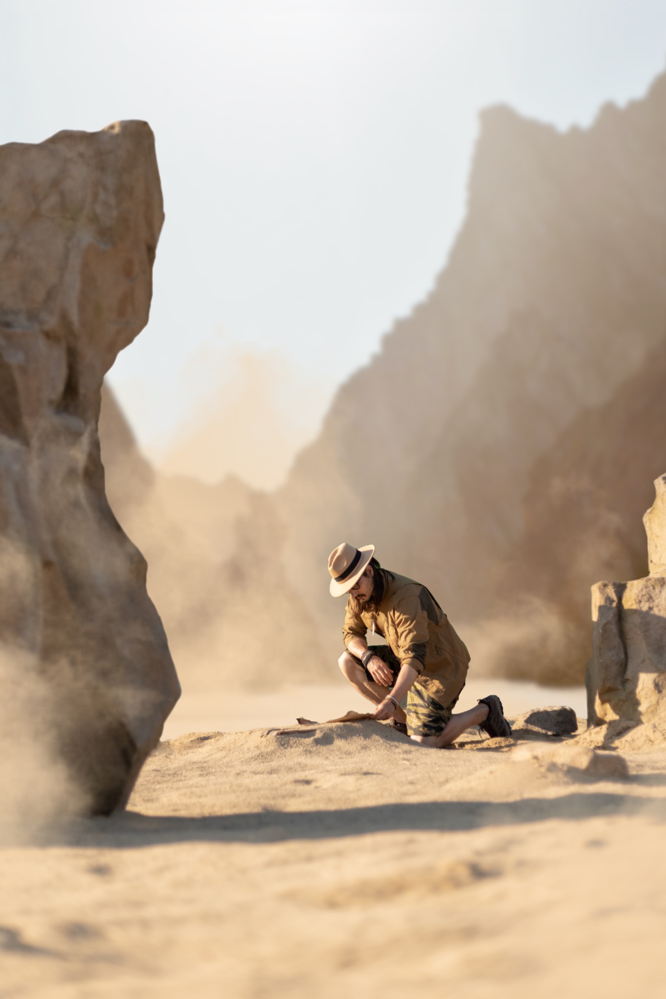 Mężczyzna w kapeluszu i polowej koszuli klęczy na piasku w pustynnym krajobrazie, badając coś na ziemi; w tle skaliste góry i unoszący się piasek.