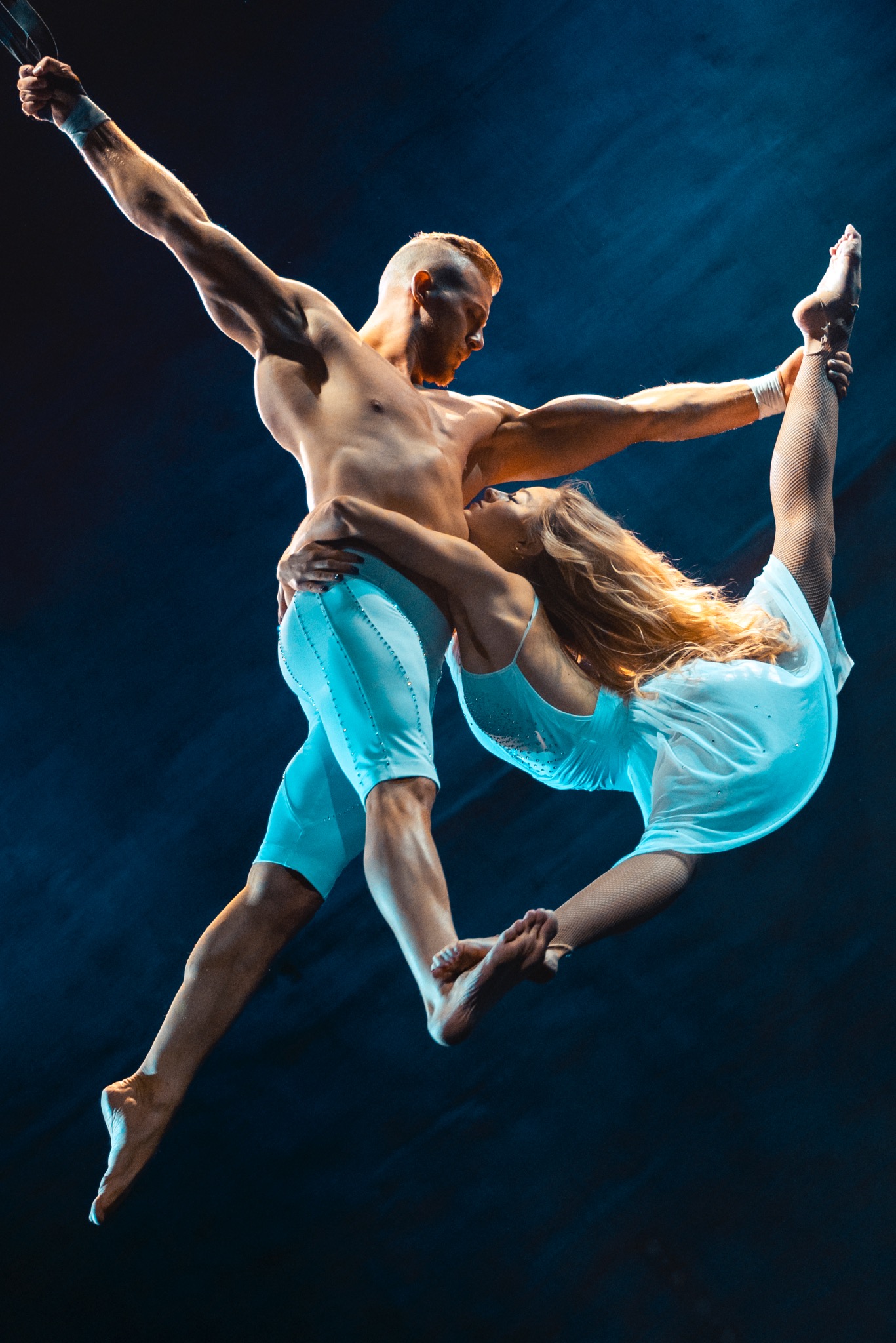 Para akrobatów w dynamicznej pozie: mężczyzna podtrzymuje kobietę, która wykonuje szpagat w powietrzu, oboje ubrani w jasne stroje na ciemnym tle.