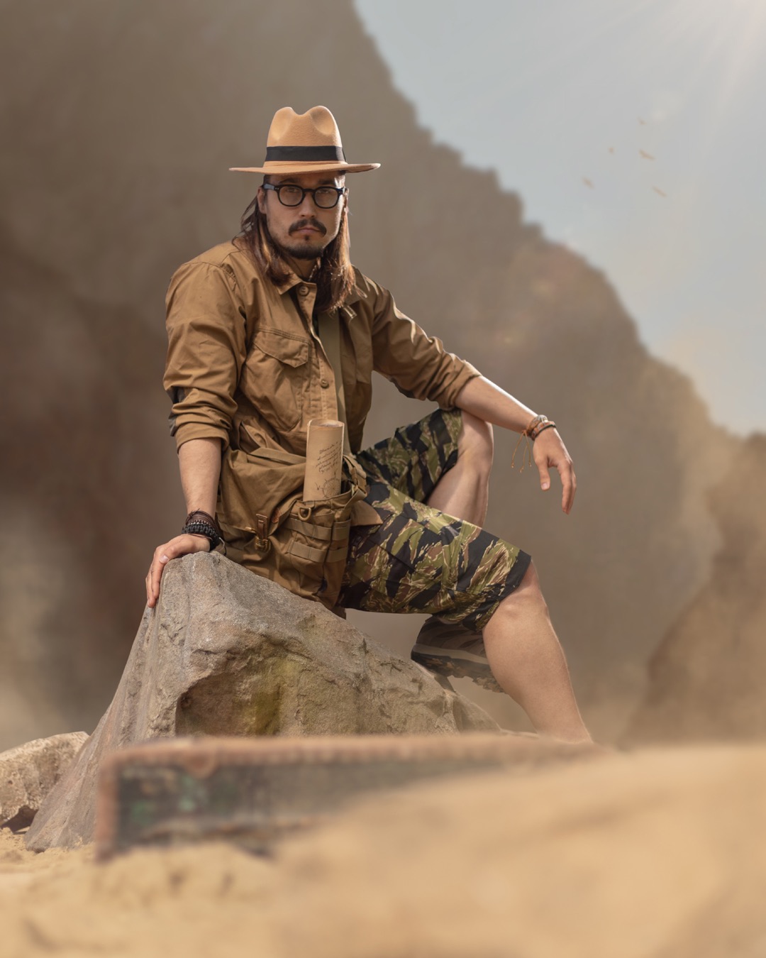 Mężczyzna w kapeluszu i okularach siedzi na kamieniu w plenerze, ubrany w koszulę khaki i szorty moro, z mapą w kieszeni; fotomanipulacja.