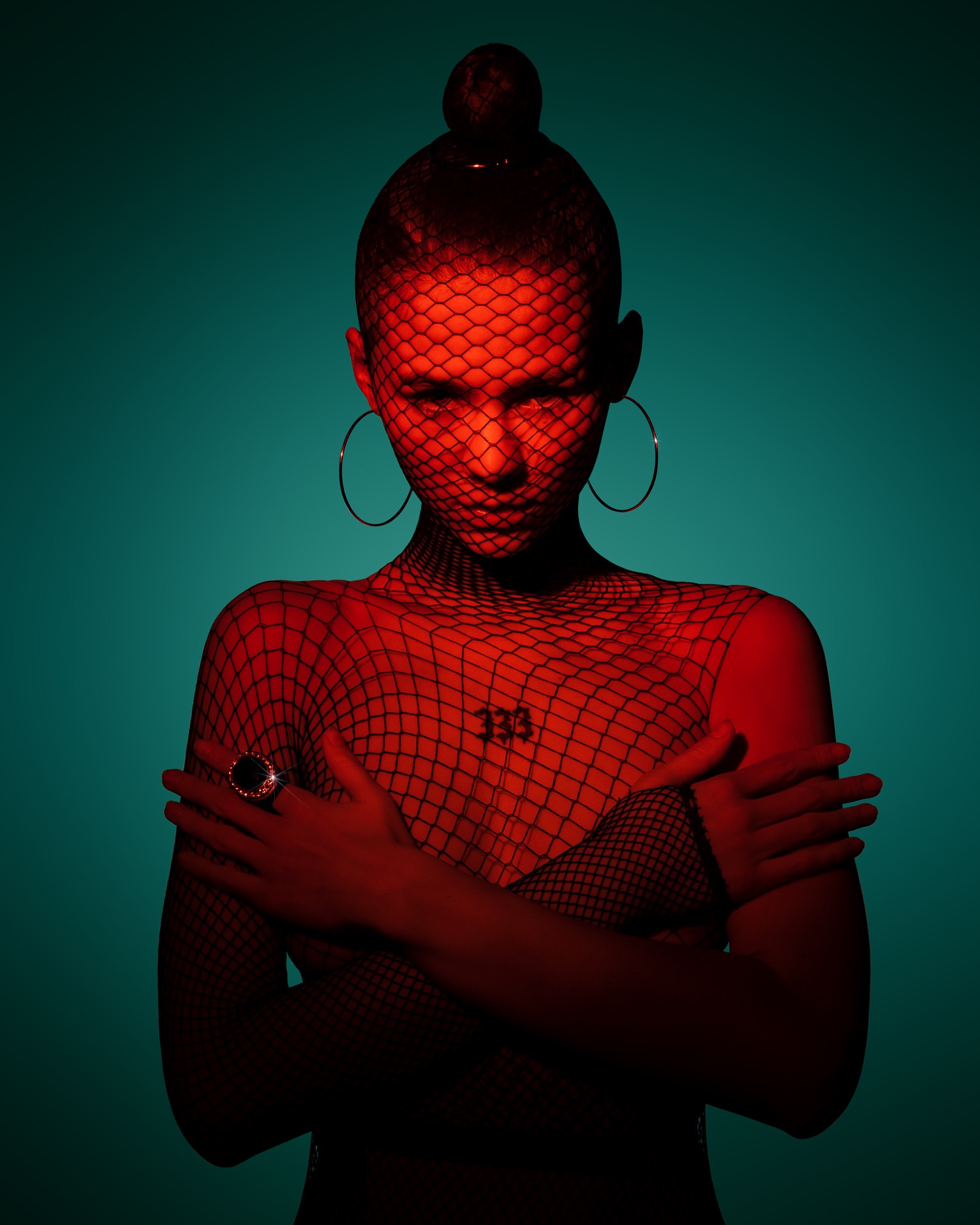 Portret kobiety z siatką na twarzy, oświetlonej czerwonym światłem, na turkusowym tle, z rękami skrzyżowanymi na piersiach i dużym pierścieniem na palcu.