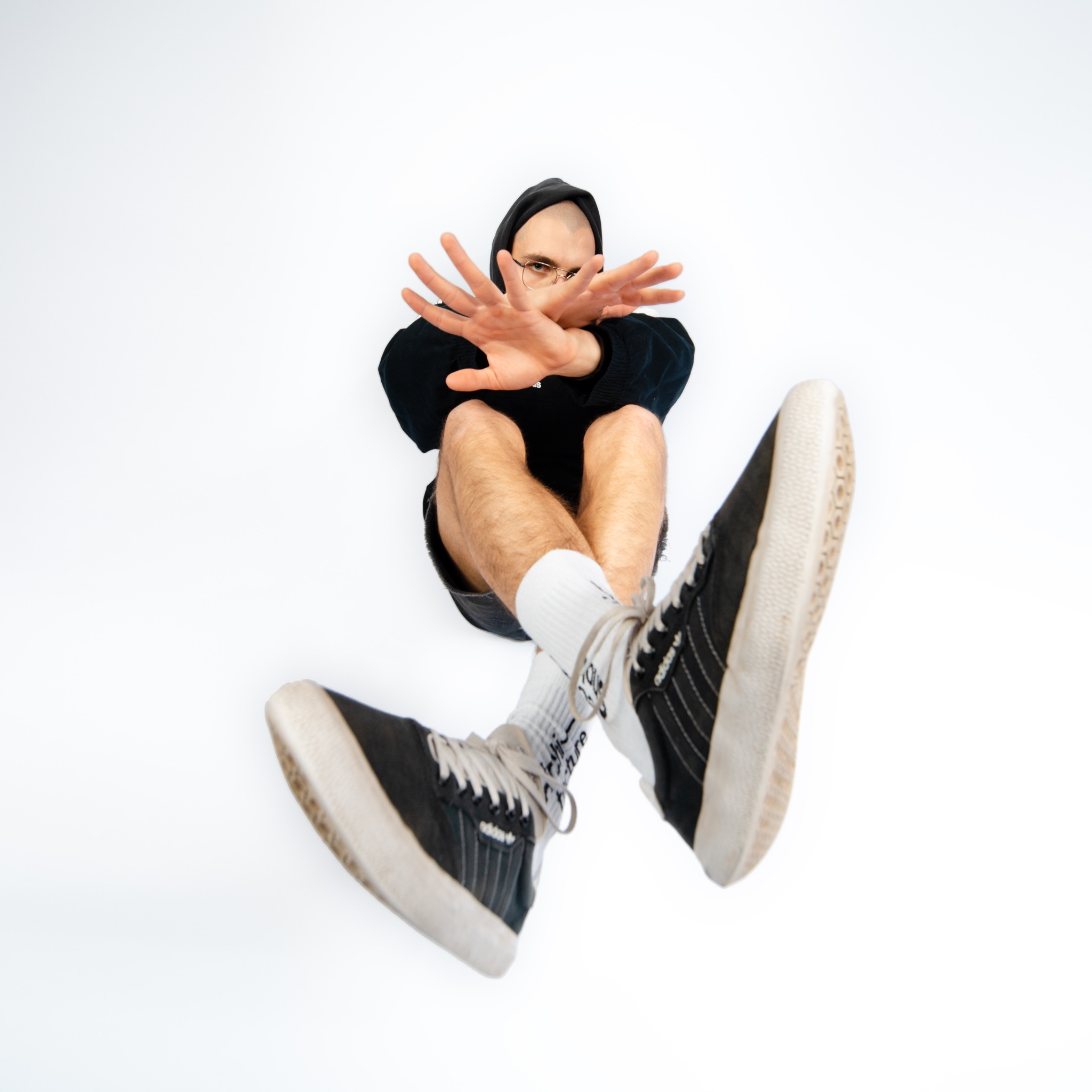 Studyjna sesja portretowa mężczyzny w czarnej bluzie z kapturem, z wyciągniętymi dłońmi w geście stop, widzianego z dołu na białym tle.