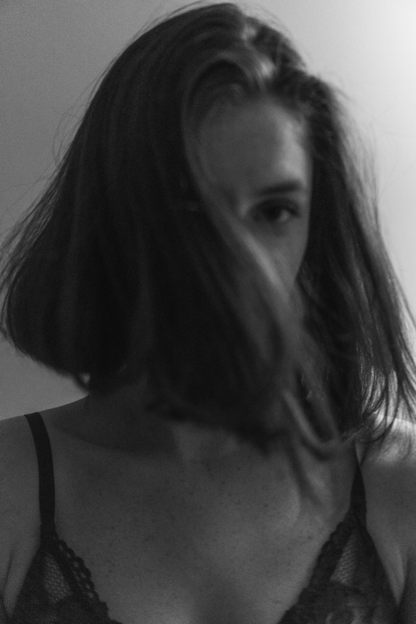 Czarno-biały portret kobiety z włosami opadającymi na twarz, widoczne ramiączka koronkowego stanika, artystyczne ujęcie z delikatnym rozmyciem.