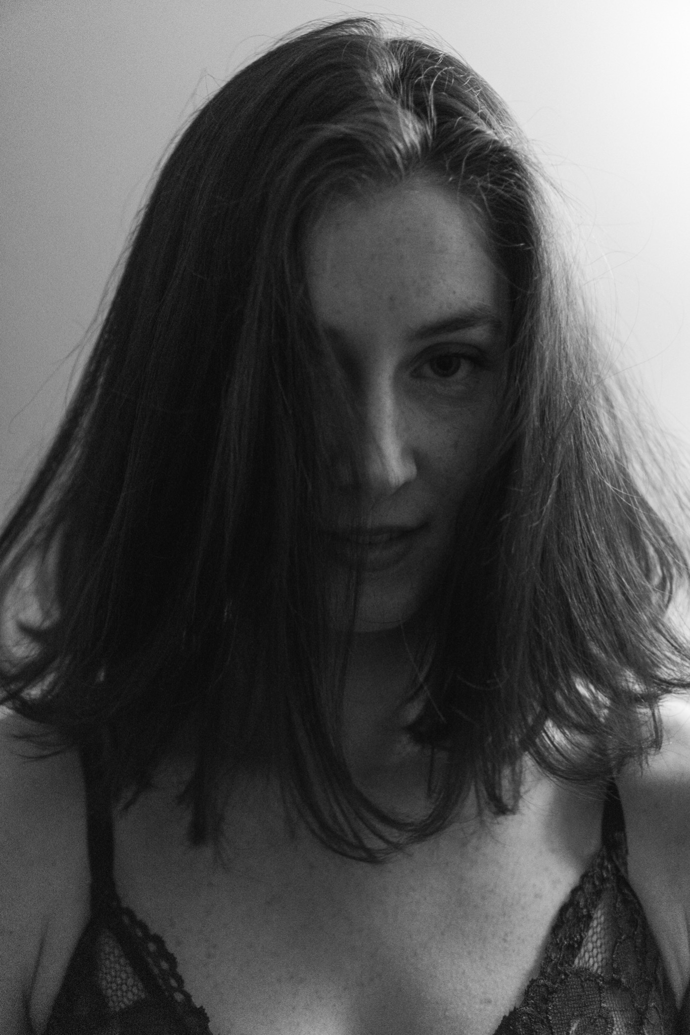 Czarno-białe, portretowe ujęcie kobiety z włosami częściowo zakrywającymi twarz, uwydatniające jej spojrzenie i delikatny uśmiech, w koronkowym staniku.