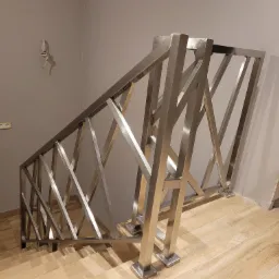 Nowoczesna, metalowa konstrukcja schodów z krzyżującymi się elementami, zamontowana na drewnopodobnych płytkach podłogowych, widoczne niedokończone wykończenie ściany.