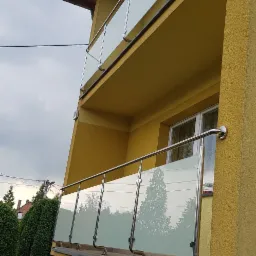 Żółty budynek z balkonami zabezpieczonymi balustradami ze szkła matowego i stali nierdzewnej, widok z dołu na tle pochmurnego nieba.