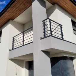 Nowoczesny balkon z czarną, minimalistyczną barierką na tle białej elewacji domu z drewnianym podsufitką.