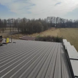 Szary dach z blachy trapezowej w trakcie montażu, widoczne narzędzia i odbicie pracownika w szybie, w tle krajobraz z drzewami i polem.