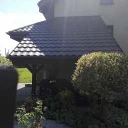 Zadaszenie tarasu z ciemną blachodachówką, widok z ogrodu z krzewami i kamieniami, fragment elewacji domu w tle.