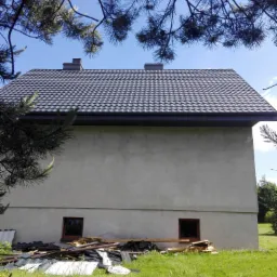 Dwuspadowy dom z szarą dach&oacute;wką, widok z dołu przez gałęzie drzewa, z gruzem budowlanym i elementami starego dachu u podstawy ściany.