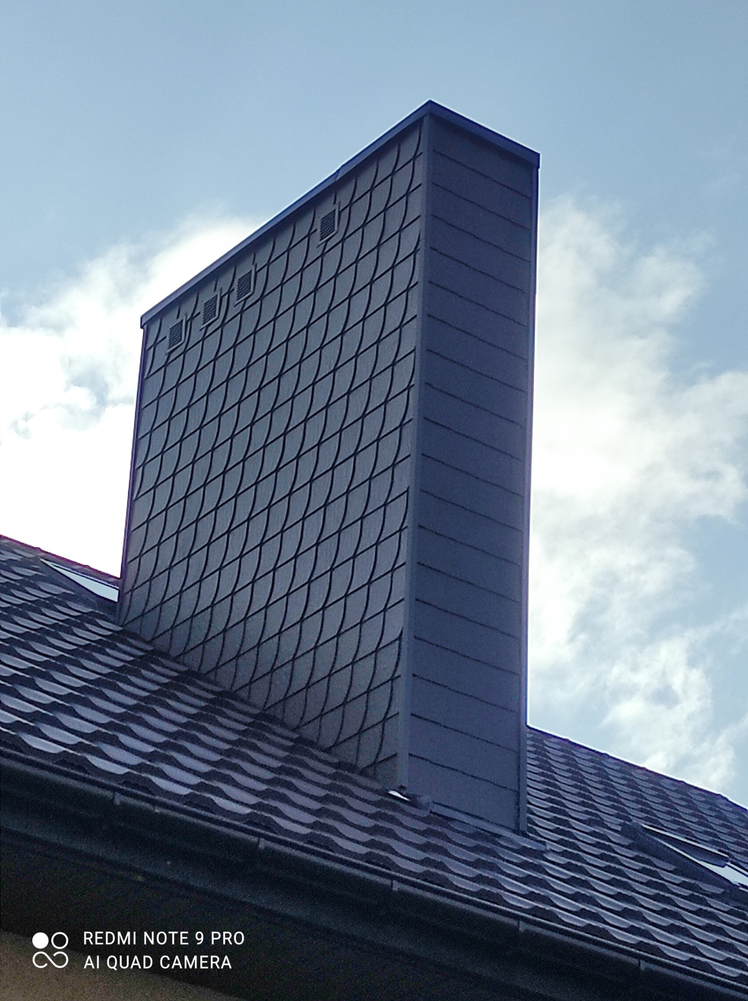 Szary komin obłożony łupkiem na tle nieba z chmurami, dopasowany do koloru dachu z dachówki bitumicznej.