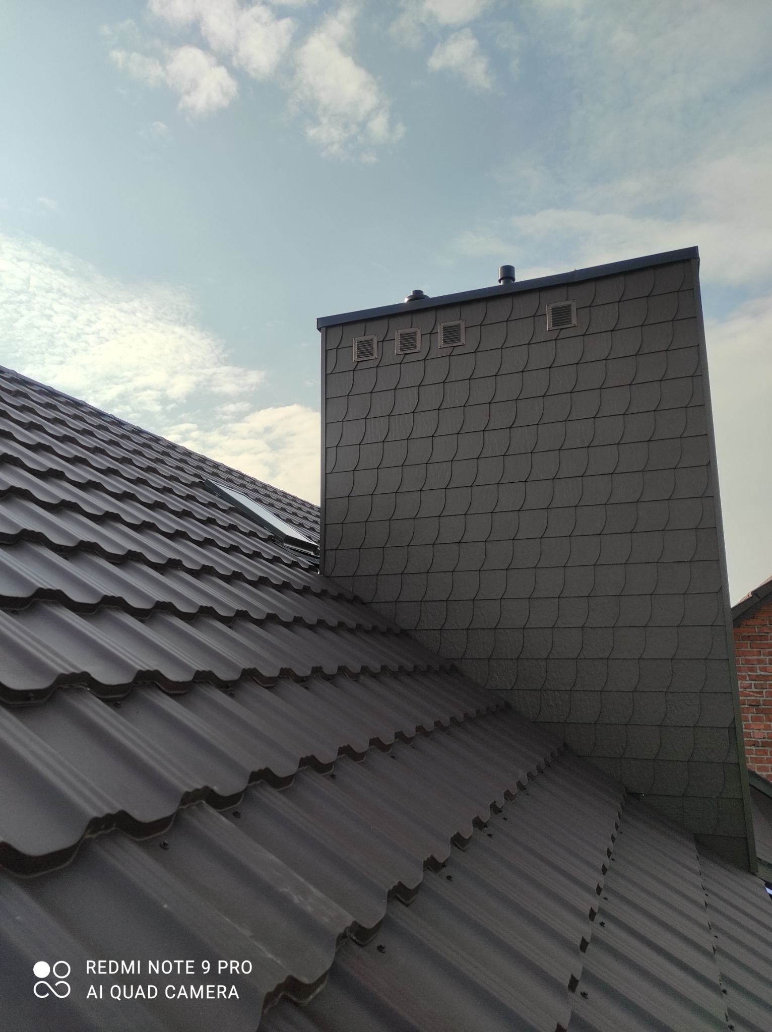 Brązowy dach z blachy trapezowej z oknem dachowym i kominem wykończonym szarą łuską na tle błękitnego nieba.