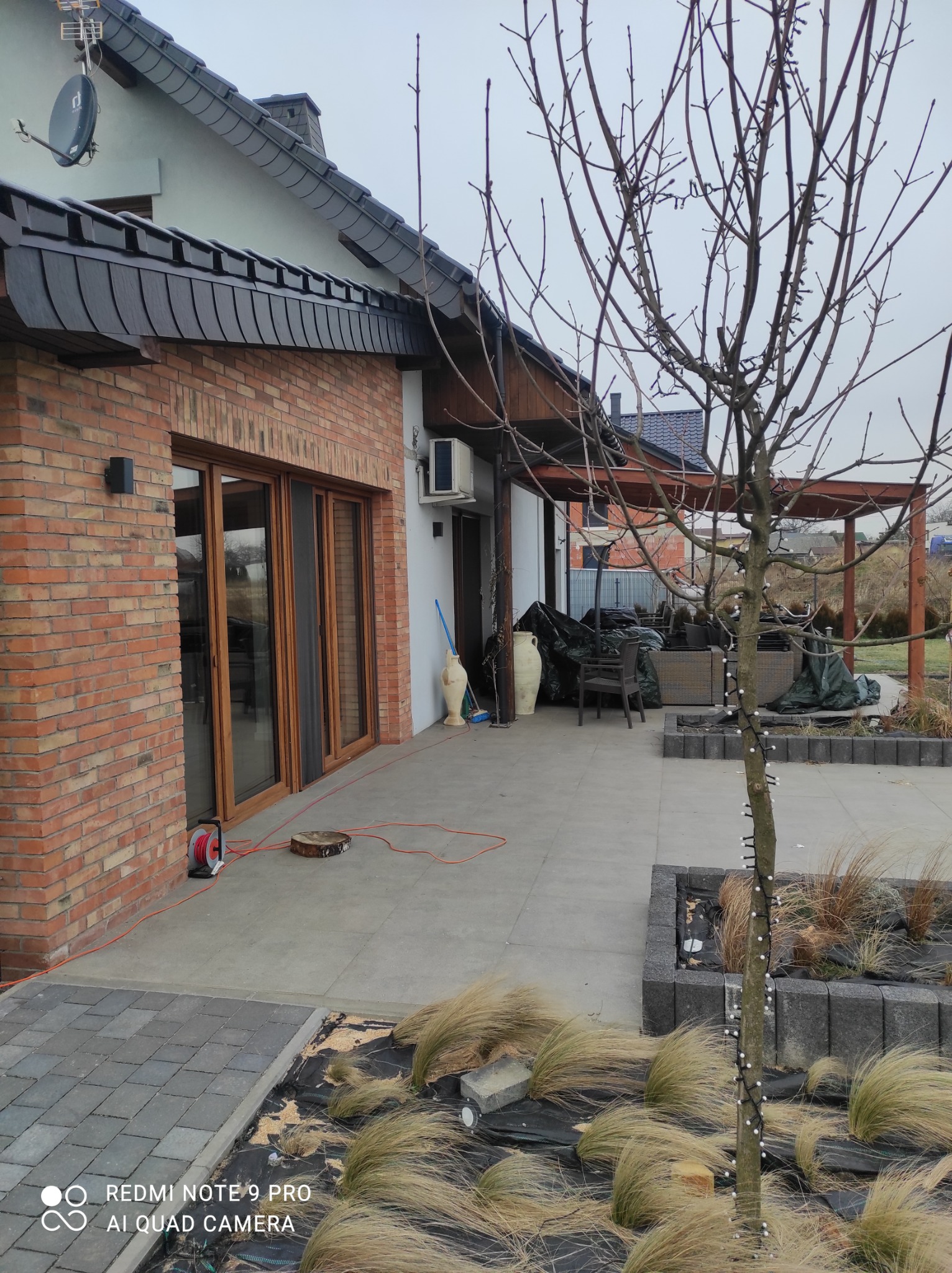 Nowoczesny dom jednorodzinny z ceglaną elewacją, dużymi przesuwnymi oknami, wykończonym dachem z ciemnej dachówki i zagospodarowanym terenem wokół, w tym patio z drewnianą pergolą i dekoracyjnym...