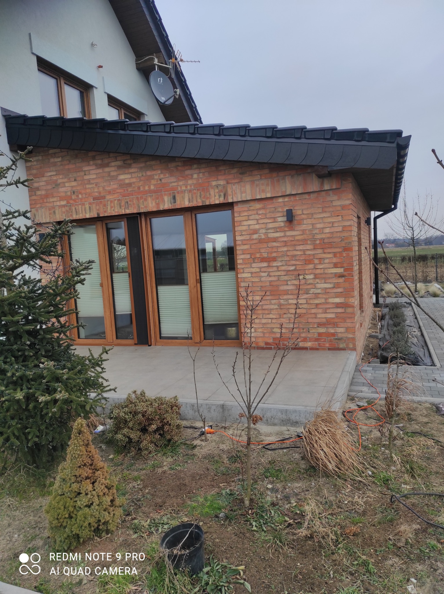 Nowoczesny, jednopiętrowy budynek z czerwonej cegły z dużymi oknami i ciemnym dachem modułowym, otoczony młodymi drzewkami i krzewami, widok z zewnątrz w pochmurny dzień.