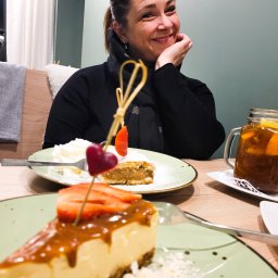 Catering Czary Mary Bożena Hanak - Uśmiechnięta kobieta siedzi przy stole z deserami: kawałek sernika z karmelem i truskawkami, ciasto z bitą śmietaną i napój w słoiku.
