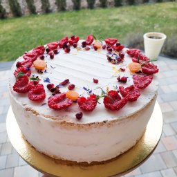 Catering Czary Mary Bożena Hanak - Tort z jasnym kremem, udekorowany malinami, granatem, physalis i suszonymi kwiatami, na złotym podkładzie, trzymany w dłoni, w tle zielony trawnik i tuje.