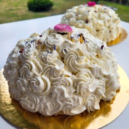 Catering Czary Mary Bożena Hanak - Dwa torty w kształcie kopuły udekorowane białym kremem w rozetki i posypane jadalnymi kwiatami, umieszczone na złotych podkładkach, z rozmytym tłem zieleni.